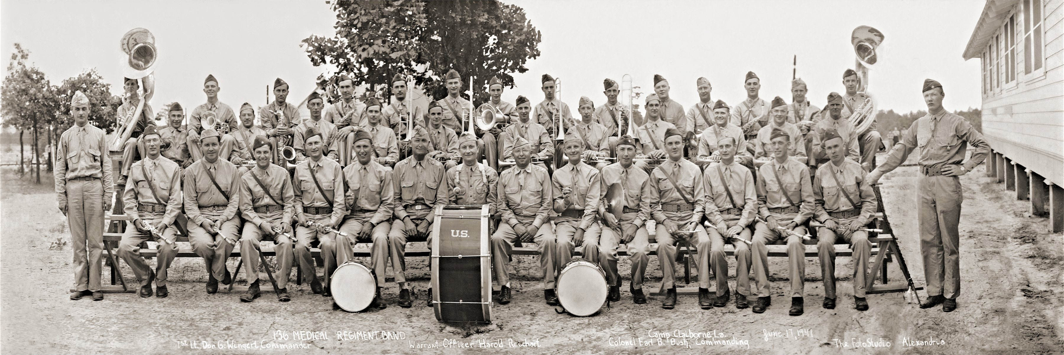 Panoramic photograph of the 136th Medical Regiment Band, some four dozen men in summer khaki with brass, woodwinds, sousaphones and two bass drums, seated and standing outside a wooden barracks at Camp Claiborne, Louisiana, June 1941.