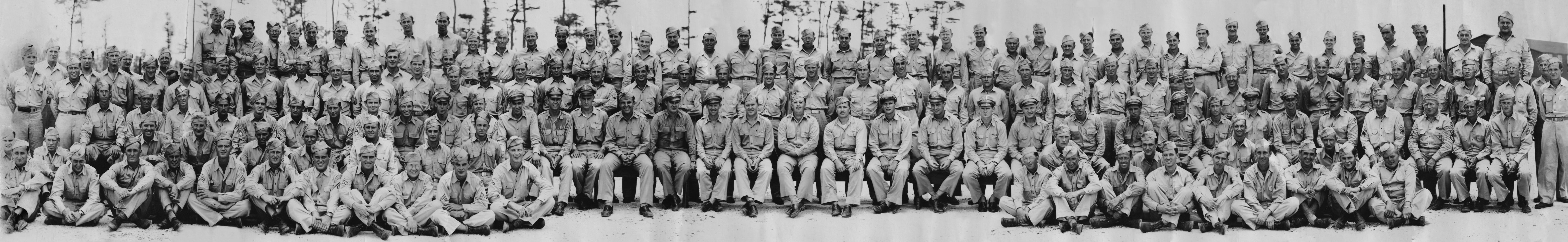 Very wide panoramic photograph of roughly two hundred airmen in summer khaki, arranged in four ranks — men seated cross-legged on sandy ground, a row seated on chairs with officers in service caps at the centre, and two standing rows behind — against a background of scrub pine.