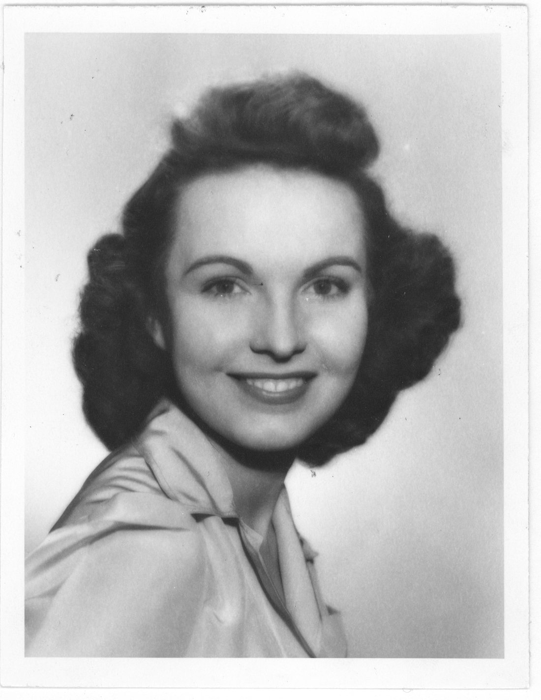 Studio portrait of a young woman with dark shoulder-length hair, smiling, in a light satin blouse with a soft bow at the collar.